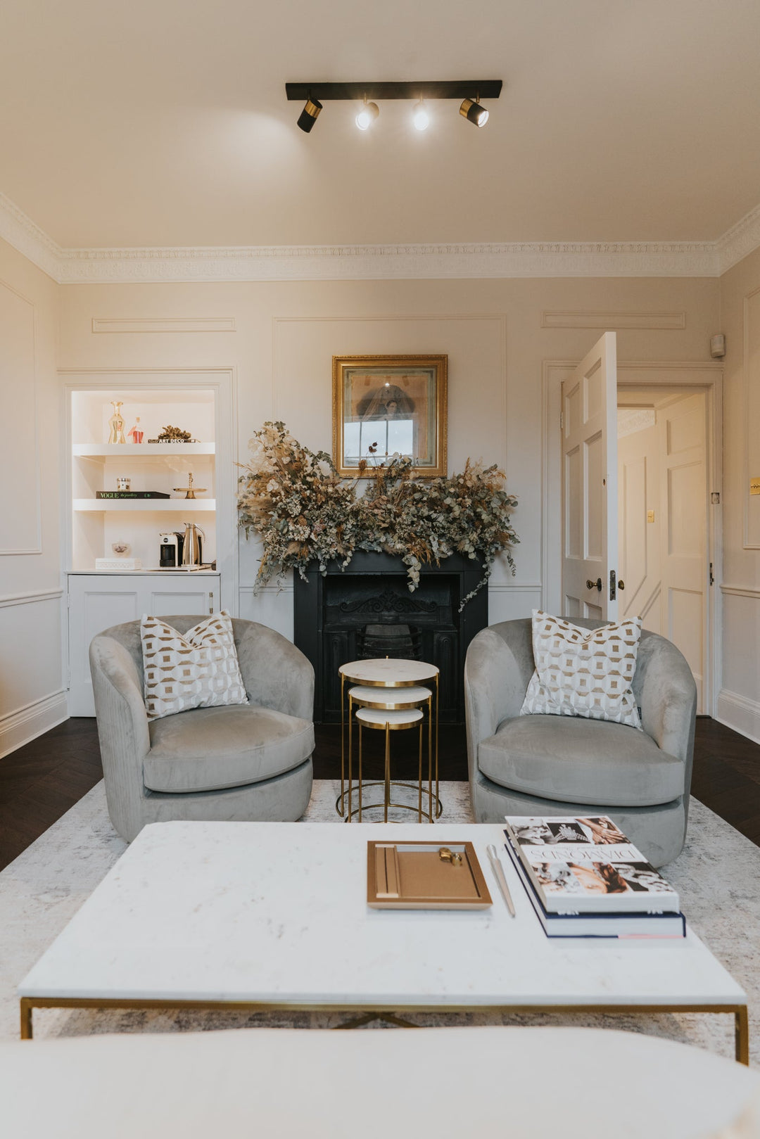 A styled interior living room scene featuring two gray armchairs with patterned pillows, a white marble coffee table with gold legs, a black fireplace with dried floral arrangements, and a track light on a cream ceiling.