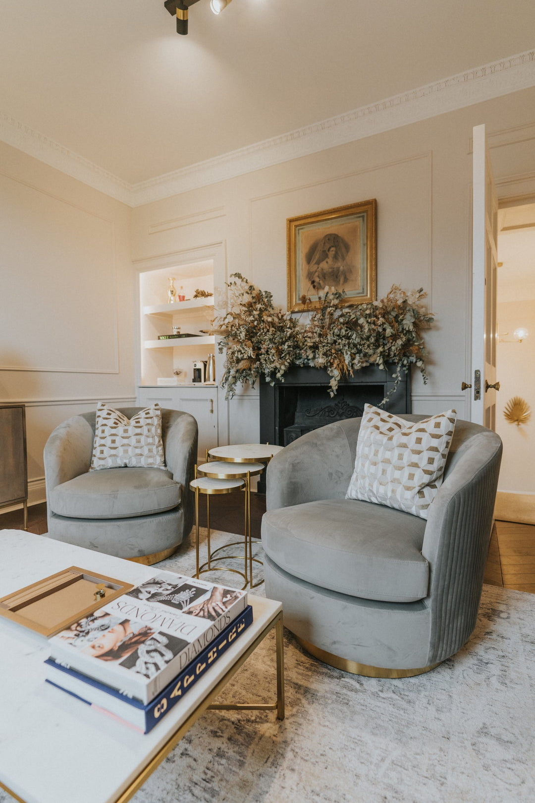 A styled interior scene showing two plush gray velvet armchairs with patterned throw pillows, a white coffee table with gold trim, a fireplace with dried floral arrangements, and a light rug in a bright, elegant room.
