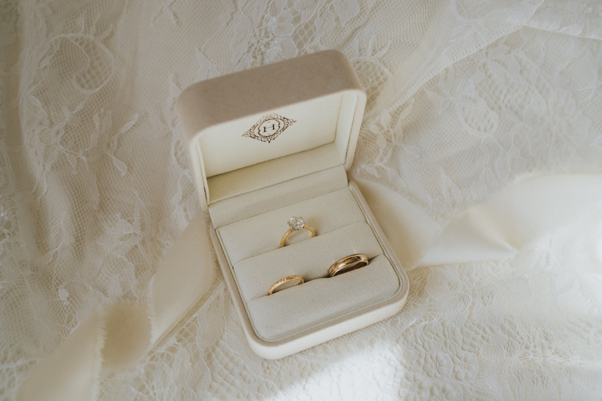 An open cream-colored velvet ring box displaying two gold wedding bands, set against a background of white lace fabric.