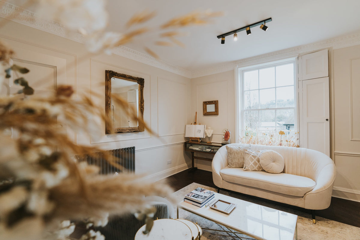 A cream-colored curved sofa in a classic living room setting, featuring a window, decorative mirror, and a blurred foreground of dried grasses.