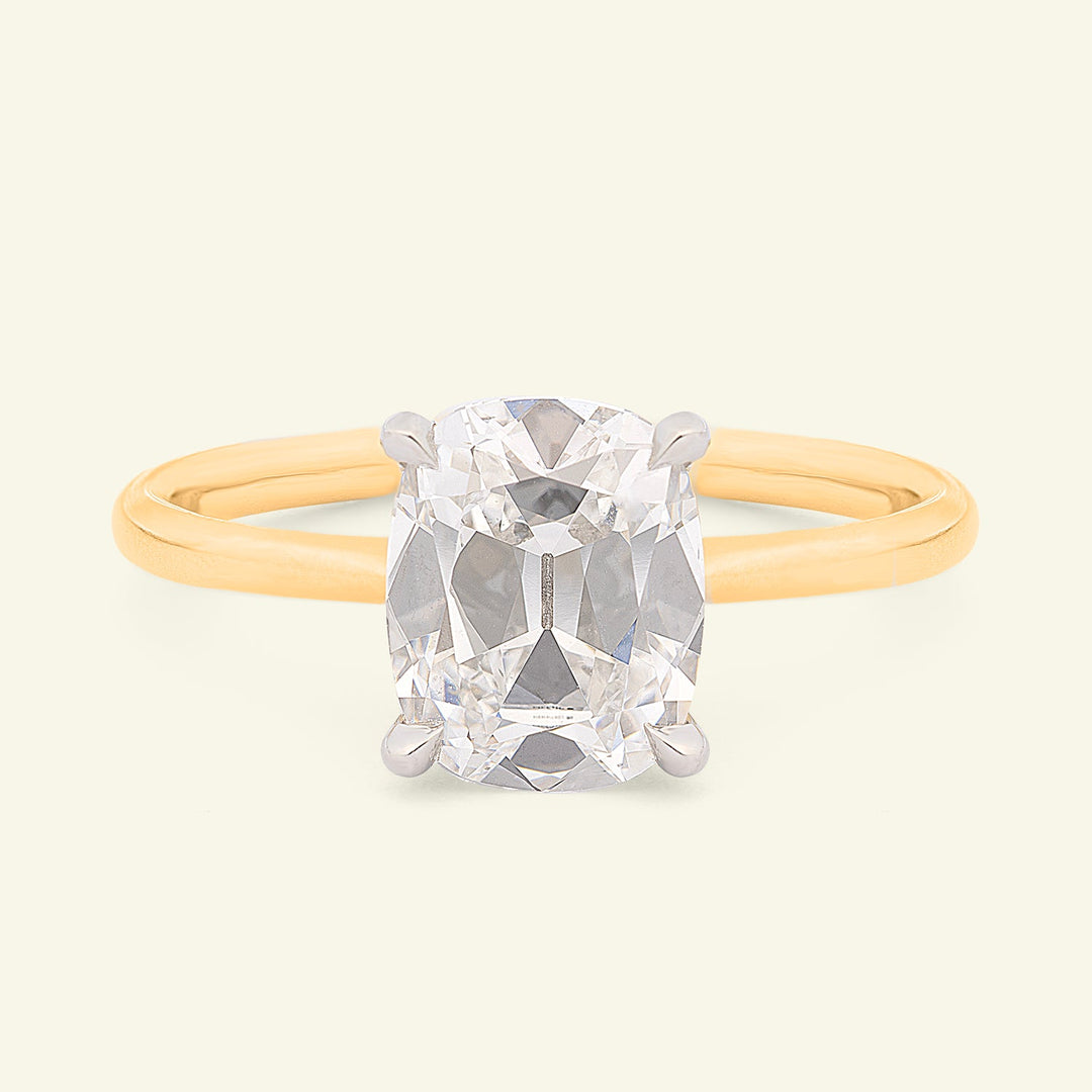 A classic solitaire engagement ring featuring a large cushion-cut diamond set in a four-prong white gold or platinum band, on a warm cream-colored background.